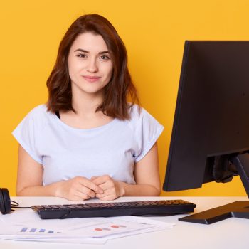 Close up portrait of beautiful young brunette female sitting at white desk in front of computer at home, has online job, isolated over yellow background, looking at camera with calm facial expression.